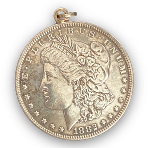 May include: A silver Morgan dollar coin pendant with a portrait of Lady Liberty on the obverse. The coin is dated 1882 and has the inscription "E Pluribus Unum" around the edge.