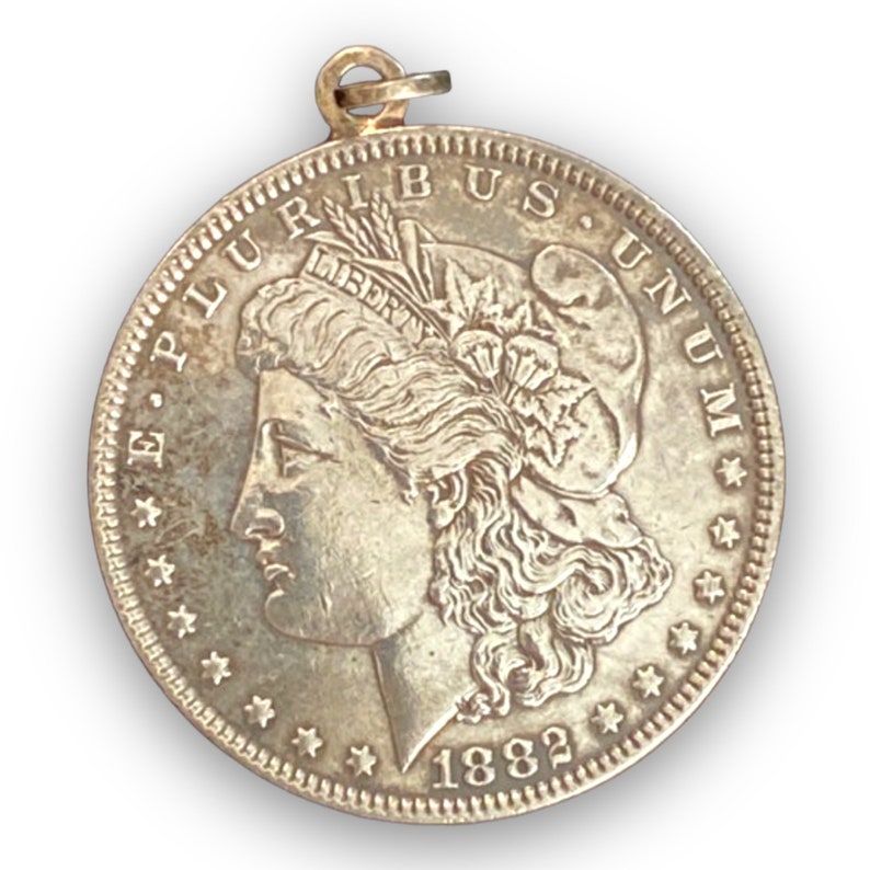 May include: A silver Morgan dollar coin pendant with a portrait of Lady Liberty on the obverse. The coin is dated 1882 and has the inscription "E Pluribus Unum" around the edge.