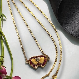 May include: A gold-toned necklace featuring a double-heart pendant. The pendant has two gold hearts, surrounded by red gemstones, and a small diamond. The necklace chain is a delicate, flat link style.