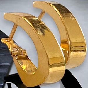 May include: A pair of gold-colored hoop earrings. The earrings have a thick, rectangular design and a hinged closure. The hoops are highly polished, reflecting light and creating a shiny appearance. The earrings are a classic jewelry item.