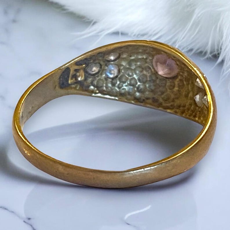 Pu&ograve; includere: Un anello d'oro con una superficie testurizzata e una pietra rosa incastonata al centro. L'anello &egrave; decorato con pietre pi&ugrave; piccole intorno al bordo.