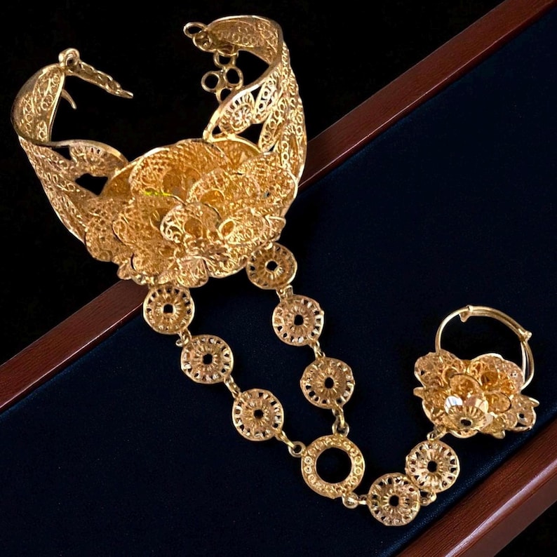May include: A gold-colored, filigree hand bracelet and ring set. The bracelet features a large floral design and connects to the ring with a chain of circular links. The jewelry is displayed on a dark blue surface.