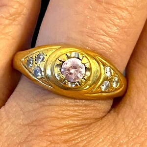 Pu&ograve; includere: Un anello d'oro con una pietra preziosa rosa circondata da pietre preziose pi&ugrave; piccole e trasparenti.