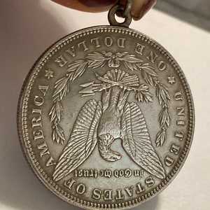 May include: Silver dollar coin pendant with a bald eagle design. The coin features the words "One Dollar", "United States of America", and "In God We Trust".