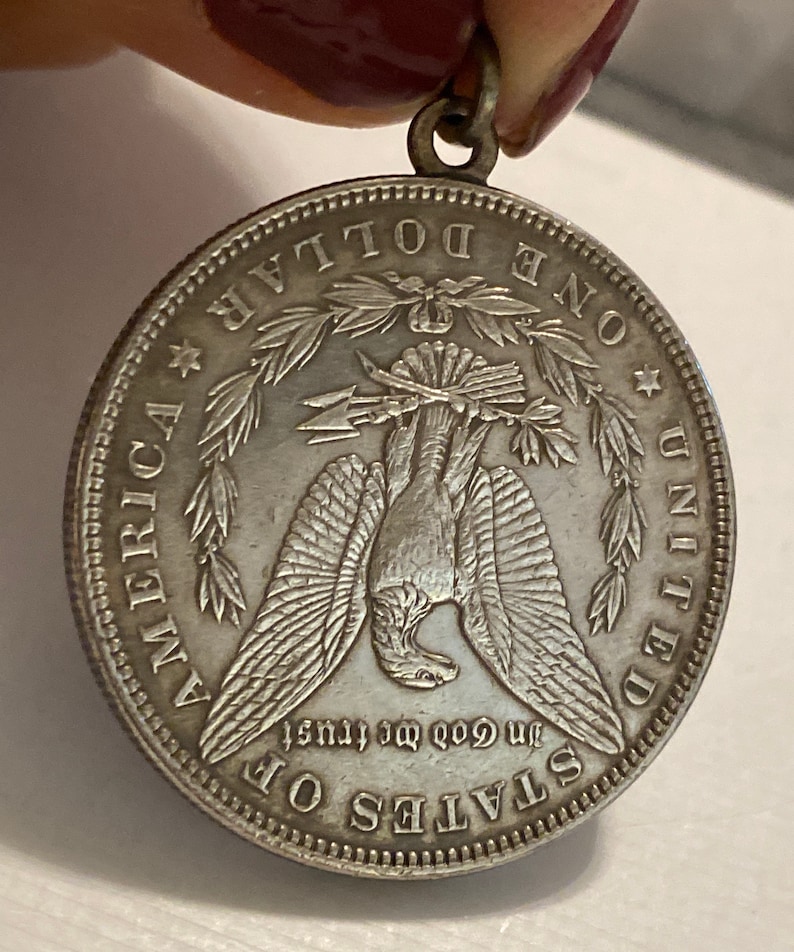May include: Silver dollar coin pendant with a bald eagle design. The coin features the words "One Dollar", "United States of America", and "In God We Trust".