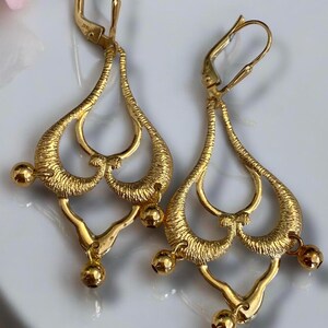 May include: A pair of gold dangle earrings with a delicate, intricate design. The earrings feature multiple loops and a dangling ball at the bottom.