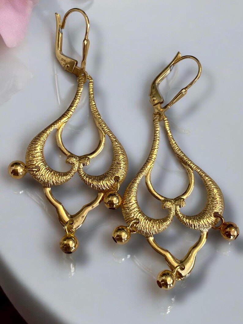 May include: A pair of gold dangle earrings with a delicate, intricate design. The earrings feature multiple loops and a dangling ball at the bottom.