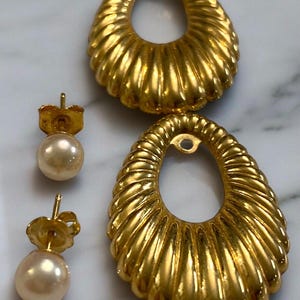 Pu&ograve; includere: Un paio di orecchini dorati con un design ovale testurizzato e un perno di perla. Gli orecchini sono su una superficie di marmo bianco.