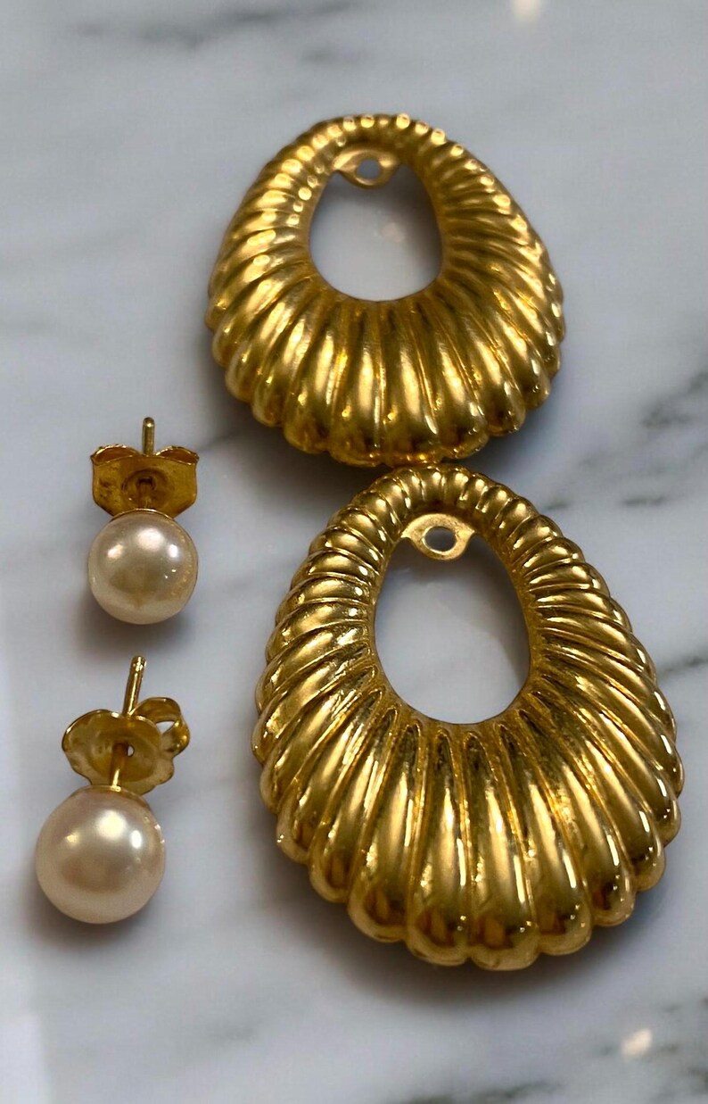 Pu&ograve; includere: Un paio di orecchini dorati con un design ovale testurizzato e un perno di perla. Gli orecchini sono su una superficie di marmo bianco.