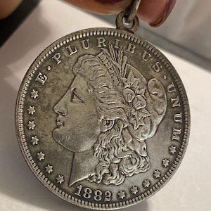 May include: A silver Morgan dollar coin with the year 1882 inscribed on the bottom. The coin features a profile of a woman with flowing hair and a wreath on her head. The inscription "E. PLURIBUS UNUM" is visible around the edge of the coin.
