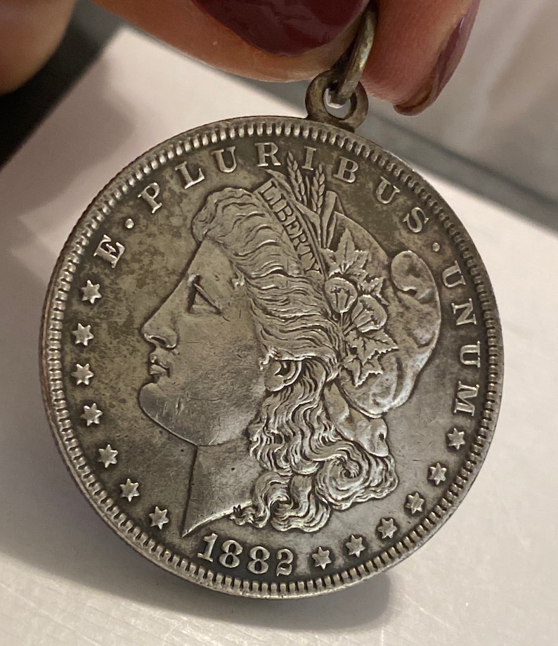 May include: A silver Morgan dollar coin with the year 1882 inscribed on the bottom. The coin features a profile of a woman with flowing hair and a wreath on her head. The inscription "E. PLURIBUS UNUM" is visible around the edge of the coin.