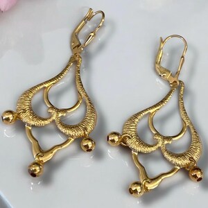 May include: A pair of gold-tone dangle earrings with a delicate, intricate design. The earrings feature multiple loops and rounded accents.