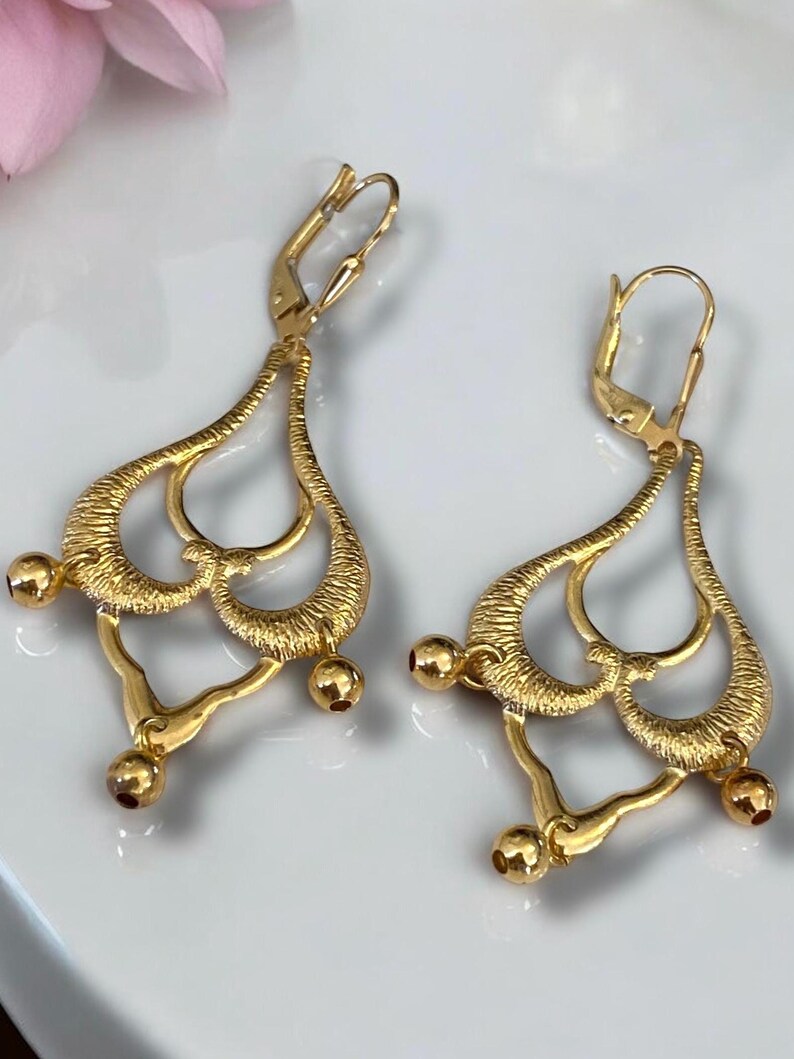 May include: A pair of gold-tone dangle earrings with a delicate, intricate design. The earrings feature multiple loops and rounded accents.