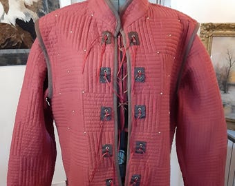 May include: A quilted, red coat with a high collar and long sleeves. The coat features a series of square patches with eyelets and red lacing down the front. Small silver studs are evenly spaced across the coat's surface. The coat is displayed on a mannequin.