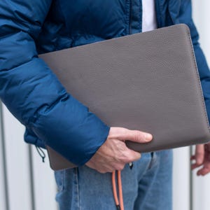 May include: A person holding a gray leather laptop sleeve. The sleeve has a textured surface and a subtle sheen.