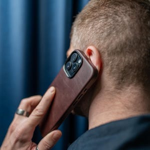 Könnte beinhalten: Eine Person hält eine braune Leder-Handyhülle mit einem schwarzen Kameraobjektiv auf einem Smartphone an ihr Ohr.