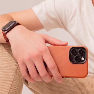 Op de afbeelding: Een persoon die een bruine leren telefoonhoes met een zwarte camera lens vasthoudt. De telefoonhoes ligt op een beige corduroy achtergrond.