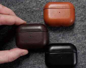 Puede incluir: Tres fundas de cuero para AirPod en marrón, marrón oscuro y negro. Las fundas son rectangulares con bordes redondeados y un pequeño agujero en la parte delantera. Las fundas están sobre una superficie gris.