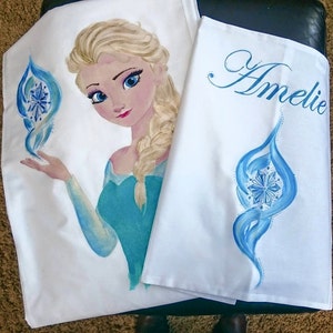 Może przedstawiać: Biała tkanina z ręcznie malowanym wizerunkiem postaci z blond włosami i niebieską sukienką. Postać trzyma niebiesko-biały wz&oacute;r. Imię "Amelie" jest napisane niebieskim pismem na drugim kawałku materiału.