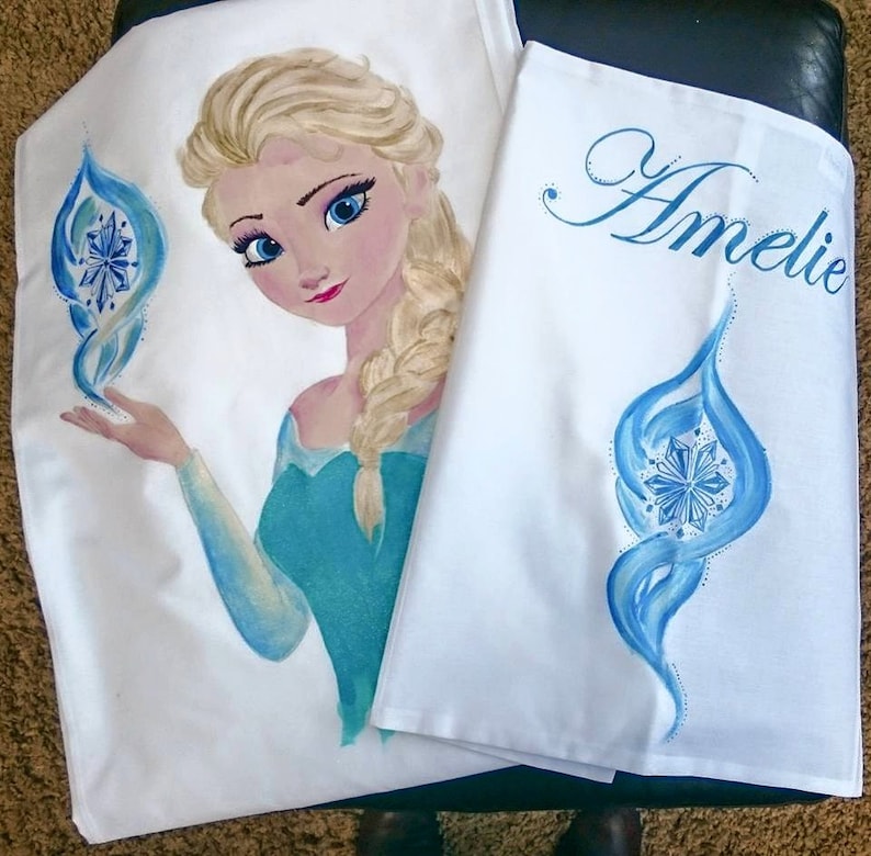 Może przedstawiać: Biała tkanina z ręcznie malowanym wizerunkiem postaci z blond włosami i niebieską sukienką. Postać trzyma niebiesko-biały wz&oacute;r. Imię "Amelie" jest napisane niebieskim pismem na drugim kawałku materiału.