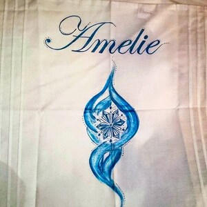 Może przedstawiać: Biała tkanina z napisem "Amelie" w niebieskim, kursywnym skrypcie. Pod imieniem znajduje się niebieski, stylizowany płomień z centralnym wzorem przypominającym płatek śniegu. Tkanina ma pionowe linie.