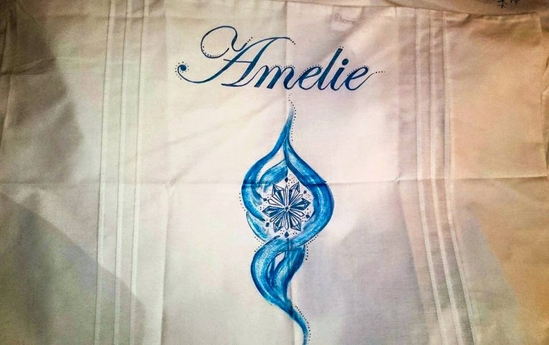 Może przedstawiać: Biała tkanina z napisem "Amelie" w niebieskim, kursywnym skrypcie. Pod imieniem znajduje się niebieski, stylizowany płomień z centralnym wzorem przypominającym płatek śniegu. Tkanina ma pionowe linie.