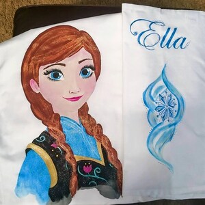 Może przedstawiać: Biała poszewka na poduszkę z ręcznie malowanym wizerunkiem postaci z kresk&oacute;wki o rudych włosach i niebieskich oczach. Poszewka ma r&oacute;wnież imię "Ella" w niebieskim skrypcie i niebieski wz&oacute;r płatka śniegu.