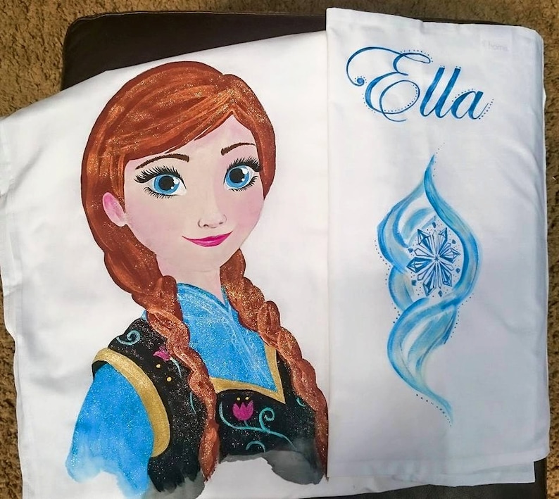 Może przedstawiać: Biała poszewka na poduszkę z ręcznie malowanym wizerunkiem postaci z kresk&oacute;wki o rudych włosach i niebieskich oczach. Poszewka ma r&oacute;wnież imię "Ella" w niebieskim skrypcie i niebieski wz&oacute;r płatka śniegu.