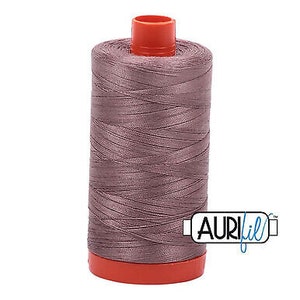 Pode incluir: Uma bobina de fio de cor malva com uma bobina de plástico vermelha. O fio é etiquetado com "Aurifil" em azul.