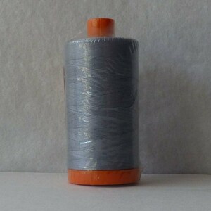 Puede incluir: Una bobina de hilo gris claro envuelta en plástico transparente con parte superior e inferior naranja.