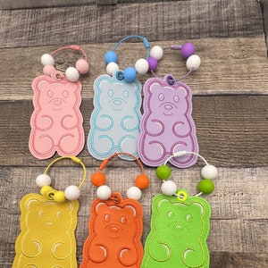 May include: A collection of colorful gummy bear-shaped baby teethers. Each teether is a different color, including pink, blue, purple, yellow, orange, and green. They have a beaded loop with white and colored beads.