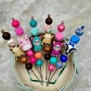 May include: A set of five decorative pens featuring colorful silicone beads and character charms including a unicorn, a cow, and dogs in hoodies. The beads are in various shapes and colors, such as pink, blue, brown, and floral patterns, arranged on metal shafts.