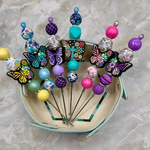 May include: A set of five metal pens decorated with colorful beads and butterfly-shaped charms. The beads feature various patterns including floral, checkered, and solid colors in shades of yellow, blue, pink, and purple. Each pen has a pointed tip.