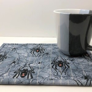 May include: A gray and white spiderweb-patterned coaster with black spiders and red accents. A black, gray, and white mug rests on the coaster. The mug has a handle and a vertical color block design.