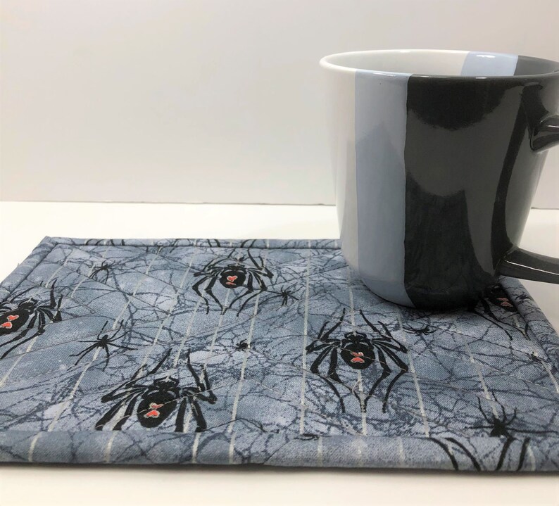 May include: A gray and white spiderweb-patterned coaster with black spiders and red accents. A black, gray, and white mug rests on the coaster. The mug has a handle and a vertical color block design.