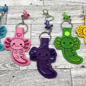 May include: Five colorful keychains featuring axolotl designs. Each keychain has a different color and features a cute axolotl with a heart on its chest. The keychains are made of felt and are attached to metal key rings.