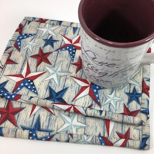 May include: A set of three red, white, and blue star-patterned fabric coasters. The coasters are layered on top of each other and feature a woodgrain background. A mug with a maroon interior and white cursive writing is sitting on top of the coasters.