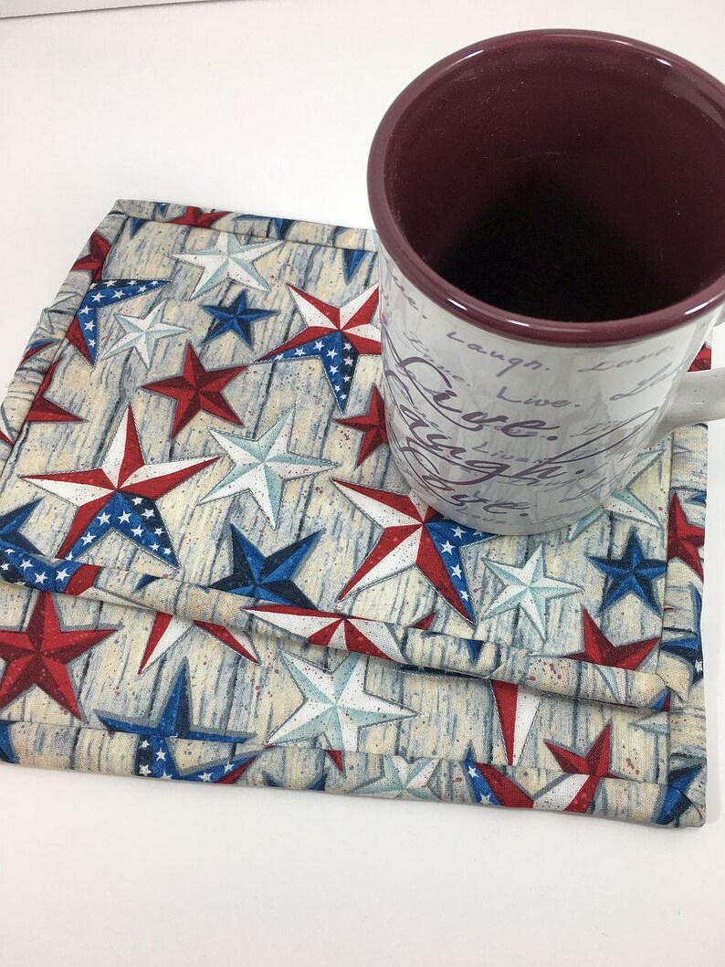 May include: A set of three red, white, and blue star-patterned fabric coasters. The coasters are layered on top of each other and feature a woodgrain background. A mug with a maroon interior and white cursive writing is sitting on top of the coasters.