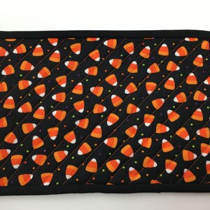 May include: A rectangular black placemat with a repeating pattern of orange, white, and yellow candy corn. The placemat has a black border and is quilted with diagonal stitching. The design is perfect for Halloween.