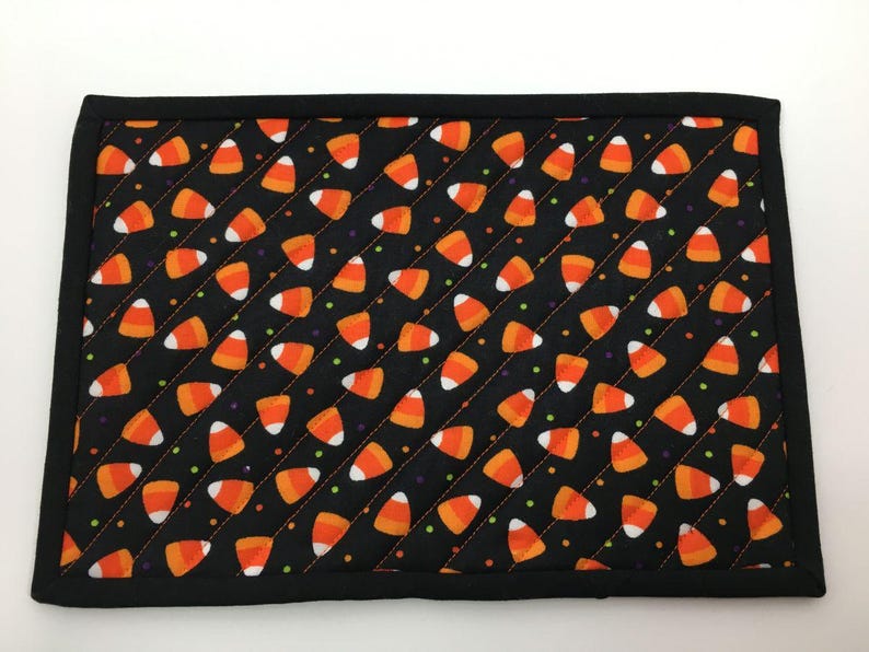 May include: A rectangular black placemat with a repeating pattern of orange, white, and yellow candy corn. The placemat has a black border and is quilted with diagonal stitching. The design is perfect for Halloween.