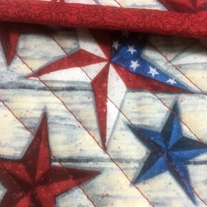 May include: A close-up of a fabric with a pattern of red, white, and blue stars on a white background. The fabric is quilted and has a subtle woodgrain texture.