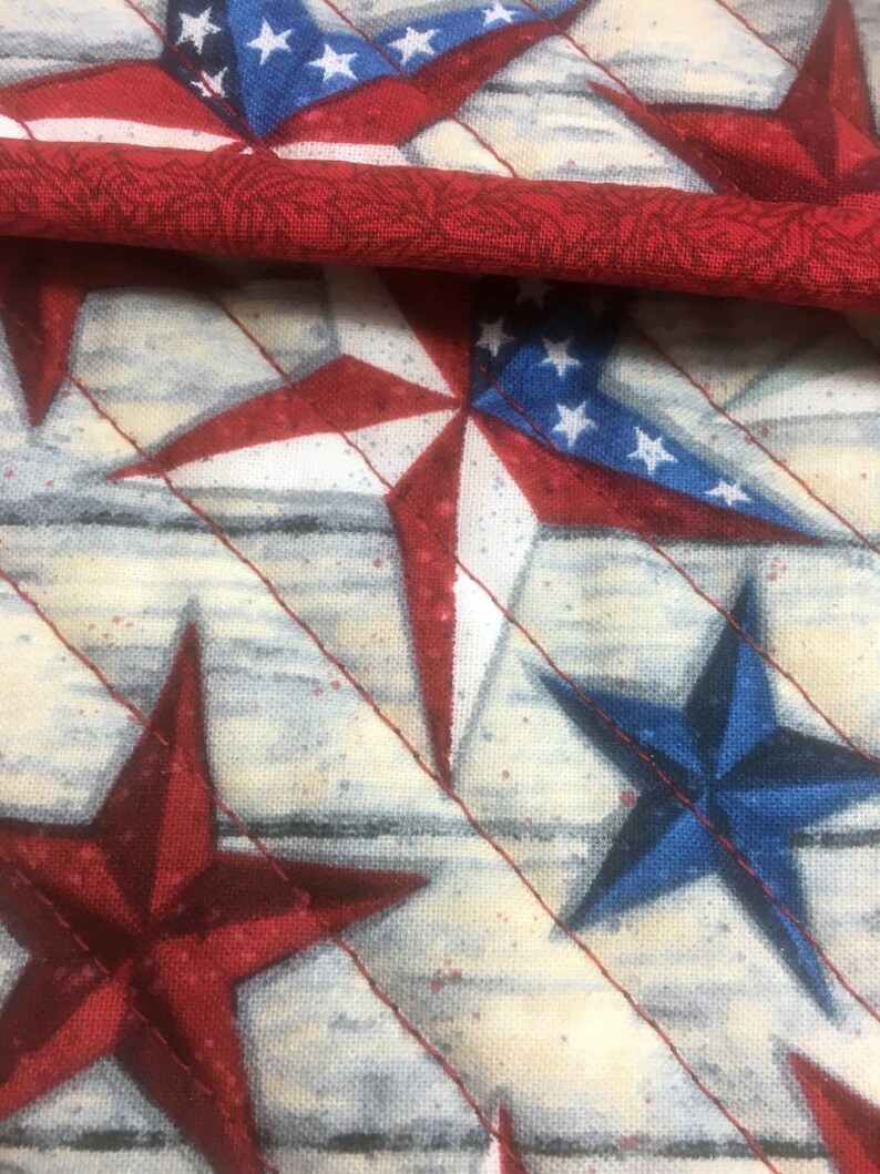 May include: A close-up of a fabric with a pattern of red, white, and blue stars on a white background. The fabric is quilted and has a subtle woodgrain texture.