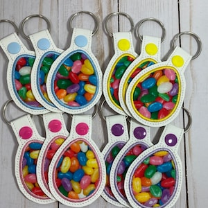 May include: Eight keychains with colorful jelly bean images inside oval-shaped frames. The keychains are made of white leather with different colored stitching around the edges. Each keychain has a different colored button.