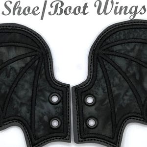 May include: A pair of black bat wings with a black and grey camouflage pattern. The wings have a stitched design and eyelets for attaching to shoes or boots.