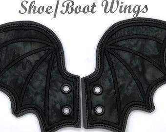 May include: A pair of black bat wings with a black and grey camouflage pattern. The wings have a stitched design and eyelets for attaching to shoes or boots.