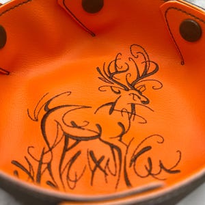 May include: An orange leather catchall tray with a brown border and snap closures. The interior features a black line drawing of a deer with antlers and grass. The tray is designed to hold small items.