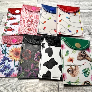 May include: A collection of small, decorative card holders with various patterns. Designs include Christmas trucks, candy canes, string lights, sunflowers, floral, cow print, and sloths. Each holder has a contrasting flap and button closure, with a variety of colors.