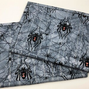 May include: Two rectangular placemats with a gray spiderweb and spider pattern. The spiders are black with red heart-shaped markings. The placemats have a quilted texture and a gray and white color scheme.