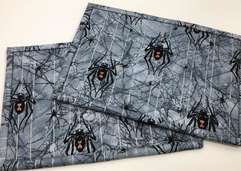 May include: Two rectangular placemats with a gray spiderweb and spider pattern. The spiders are black with red heart-shaped markings. The placemats have a quilted texture and a gray and white color scheme.