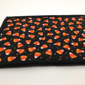 May include: A black quilted placemat featuring a pattern of orange, white, and yellow candy corn. The placemat has a black border and is decorated with small dots of green, purple, and yellow.